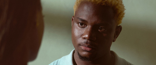 Ein nachdenklicher Mann mit blond gefärbten Haaren schaut eine Person an, deren Gesicht nicht sichtbar ist. Hintergrund unscharf.