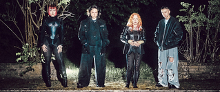 Vier Personen in schwarzer Kleidung stehen nebeneinander vor einem dunklen, bewachsenen Hintergrund. Beleuchtung hebt sie hervor.