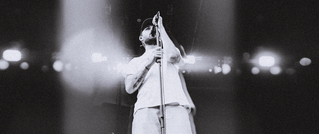 Ein Sänger steht auf einer Bühne, singt in ein Mikrofon und ist in grelles Licht getaucht. Die Szene ist schwarz-weiß.