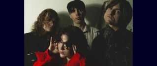 Vier Menschen stehen zusammen vor einer Wand, die Person vorne trägt eine rote Jacke und hebt die Hände zum Gesicht.