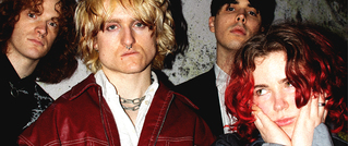 Vier Personen mit unterschiedlichen Frisuren und Outfits stehen vor einer betonierten Wand. Sie schauen ernst, eine Person stützt das Gesicht in die Hände.