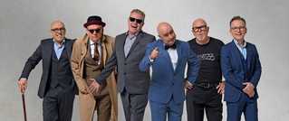Sechs Männer in Anzügen posieren lächelnd vor einem grauen Hintergrund. Einer trägt einen Hut und zwei haben Brillen.