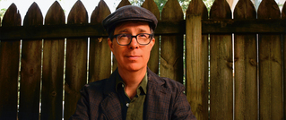 Eine Person mit Brille und Mütze steht vor einem hölzernen Zaun. Sie trägt ein kariertes Sakko und ein Hemd.