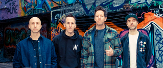 Vier Männer stehen vor einer Wand mit bunter Graffiti. Einer hebt den Daumen. Alle tragen legere Kleidung.