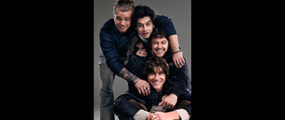 Vier Männer posieren fröhlich übereinander gestapelt. Sie lachen und tragen legere Kleidung vor einem grauen Hintergrund.