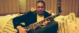 Ein Mann sitzt auf einem Sofa mit gestreiften Kissen und hält ein Saxophon in den Händen, umgeben von warmer Beleuchtung.