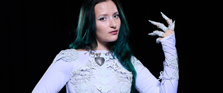 Eine Person mit langen, grünen Haaren trägt ein weißes, verziertes Outfit und zeigt kunstvoll verlängerte Fingernägel.