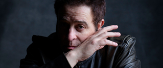 Eine Person in einer schwarzen Lederjacke schaut nachdenklich, die Hand teilweise vor das Gesicht gehalten, vor dunklem Hintergrund.