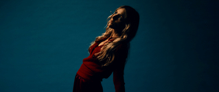 Eine Person in roter Kleidung lehnt sich in einem dunklen, stimmungsvollen Licht gegen einen blauen Hintergrund.