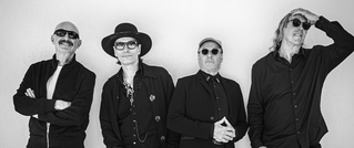 Vier Männer in schwarzer Kleidung, teils mit Sonnenbrillen und Hüten, posieren vor einer weißen Wand. Einer hält sich die Hand an die Stirn.