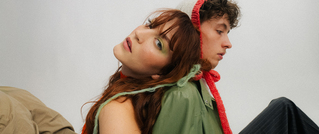 Ein Mann und eine Frau sitzen Rücken an Rücken. Die Frau trägt grünes Augen-Make-up und der Mann einen roten Schal.