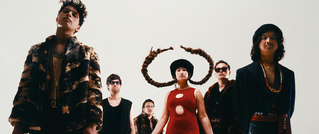 Sechs Jugendliche in auffälliger Kleidung und Hüten posieren vor weißem Hintergrund. Eine Person trägt große, skulpturale Haarhörner.