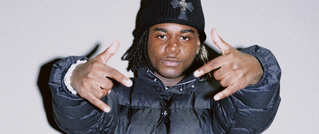 Eine Person in dunkler Winterjacke und Mütze, die beide Hände mit gespreizten Fingern gestikuliert. Hintergrund ist neutral.