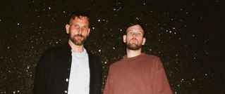 Zwei Männer stehen vor einem funkelnden, dunklen Hintergrund. Der linke trägt eine Jacke, der rechte einen Pullover.