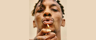 Person mit geflochtenem Haar hält einen brennenden Streichholz nahe am Gesicht und pustet darauf. Hintergrund ist einfarbig beige.