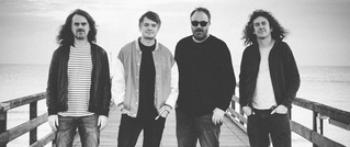 Vier Männer stehen nebeneinander auf einem Steg am Meer, in schwarz-weiß aufgenommen. Alle schauen in die Kamera.