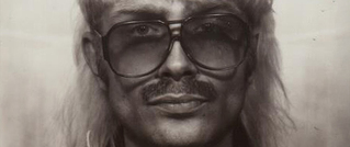 Porträt eines Mannes mit Schnurrbart, großer Brille und hellem, langem Haar, das nach hinten gekämmt ist. Das Bild ist schwarz-weiß.