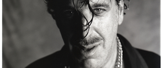 Schwarz-weiß-Foto eines Mannes mit nassen Haaren im Gesicht, Bart und einer Kette, halbes Gesicht im Schatten.