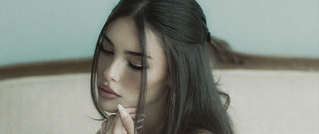 Eine Frau mit dunklen Haaren schaut nach unten, Hände nah am Gesicht. Der Hintergrund ist unscharf, die Stimmung wirkt nachdenklich.