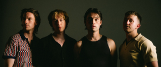 Vier Personen stehen nebeneinander und schauen in die Kamera, vor einem dunklen Hintergrund, in schummrigem Licht.