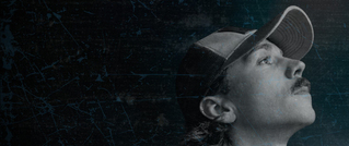 Profil eines nach oben schauenden Mannes mit Schnurrbart und Mütze, vor dunklem, verwittertem Hintergrund.