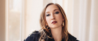 Eine Frau mit langen, blonden Haaren trägt eine schwarze Jacke vor einem Hintergrund aus weißen Vorhängen. Sie blickt ernst.