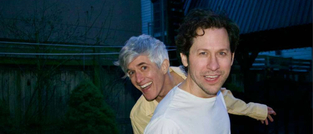 Zwei Männer stehen draußen. Der Mann vorne trägt ein weißes Shirt, der hintere ein gelbes Hemd. Beide wirken fröhlich. Hintergrund: Bäume.