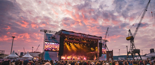 Open-Air-Konzert bei Sonnenuntergang, Bühne mit Lichtern, viele Zuschauer, Kräne und Wolken am Himmel im Hintergrund.