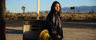 Ein Mann mit langer schwarzer Jacke steht draußen in einer Wüstenlandschaft neben einer alten Zapfsäule, eine Gitarre in der Hand.