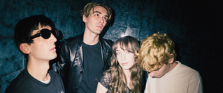 Vier Personen posieren in einem dunklen Raum. Drei blicken ernst, eine Person schaut nach unten. Einer trägt Sonnenbrillen.