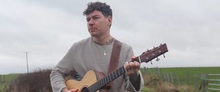 Eine Person mit Gitarre steht in einer ländlichen Umgebung, umgeben von Feldern und einem bewölkten Himmel.