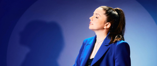 Eine Person im blauen Blazer steht seitlich, mit geschlossenen Augen und zurückgebundenem Haar, vor einem blau gemischten Hintergrund.