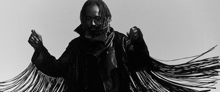 Person mit langen Dreadlocks in einer Jacke mit Fransen, erhobene Hände vor grauem Hintergrund.