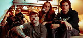 Vier Personen sitzen in einem Zimmer mit Backsteinmauer und Sofas, alle tragen legere Kleidung. Zwei haben tätowierte Arme.