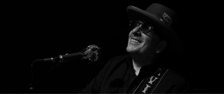 Ein Musiker mit Hut und Sonnenbrille lächelt und singt in ein Mikrofon. Das Bild ist in Schwarz-Weiß gehalten.