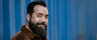 Ein lächelnder Mann mit dunklem Bart steht vor einer blauen Wand und trägt eine braune Jacke.