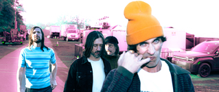 Vier Personen gehen einen Weg entlang, im Hintergrund stehen LKWs. Eine Person vorne trägt eine orangefarbene Mütze.