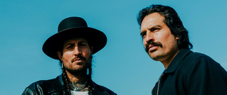 Zwei Männer vor blauem Himmel: einer mit Hut und Lederjacke, der andere mit Schnurrbart. Beide schauen ernst.