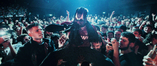 Eine Person mit Sonnenbrille tanzt inmitten einer begeisterten Menschenmenge bei einem Konzert. Viele Handys sind sichtbar.