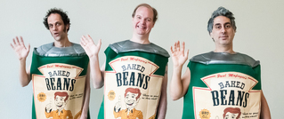 Drei Personen winken in Kostümen, die wie Baked Beans-Dosen aussehen, mit grünen und weißen Aufschriften. Sie lächeln.