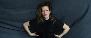 Frau mit dunklem, gewelltem Haar liegt in schwarzem Kleid auf dunkelgrauem Stoff, die Hände an den Hüften, blickt direkt zur Kamera.