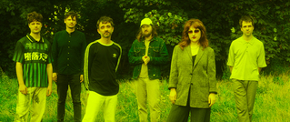 Sechs Personen stehen in einer Wiese vor einem Hintergrund aus Bäumen. Sie tragen lässige Kleidung, das Bild hat einen grünen Farbstich.