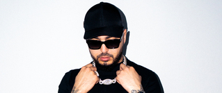 Person mit schwarzer Mütze und Sonnenbrille hält eine silberne Kette. Hintergrund ist schlicht weiß.