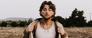 Ein Mann steht auf einem Feld, trägt ein offenes Hemd über einem weißen Tanktop und hat welliges Haar. Im Hintergrund sind Bäume.