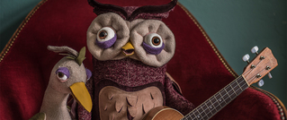 Zwei Stofftiere, eine Eule und ein Vogel, sitzen auf einem roten Sessel. Die Eule hält eine kleine Gitarre.