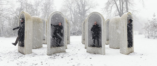 Vier Personen in schwarzen Mänteln stehen in einem verschneiten Park zwischen großen, bogenförmigen Betonplatten.