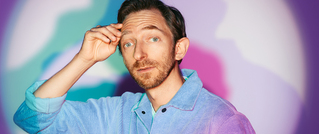 Ein Mann in einem blauen Hemd steht vor einem bunten, verschwommenen Hintergrund und berührt seine Stirn mit der Hand.