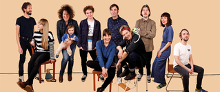 Eine Gruppe von elf Menschen, einige sitzen, andere stehen. Ein Kleinkind wird von einem Erwachsenen gehalten. Hintergrund einfarbig beige.