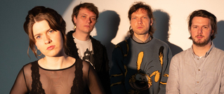 Vier Personen stehen vor einem hellen Hintergrund. Eine Person trägt ein schwarzes Netz-Oberteil, die anderen Freizeitkleidung.