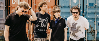 Vier Personen stehen vor bunten Schiffscontainern. Sie tragen lässige Kleidung und Sonnenbrillen und schauen in verschiedene Richtungen.